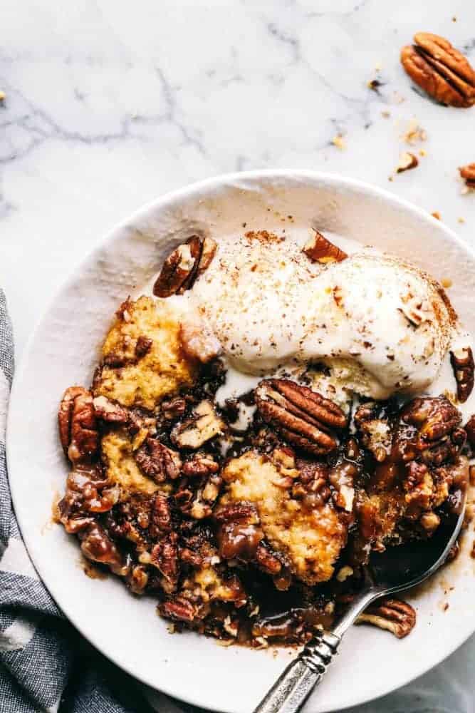 A white bowl full of Pecan Pie Cobbler with vanilla ice cream and a metal spoon. 