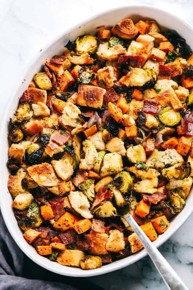 Roasted autumn vegetable stuffing on in a white baking dish with a silver spoon pulling up part of the stuffing.