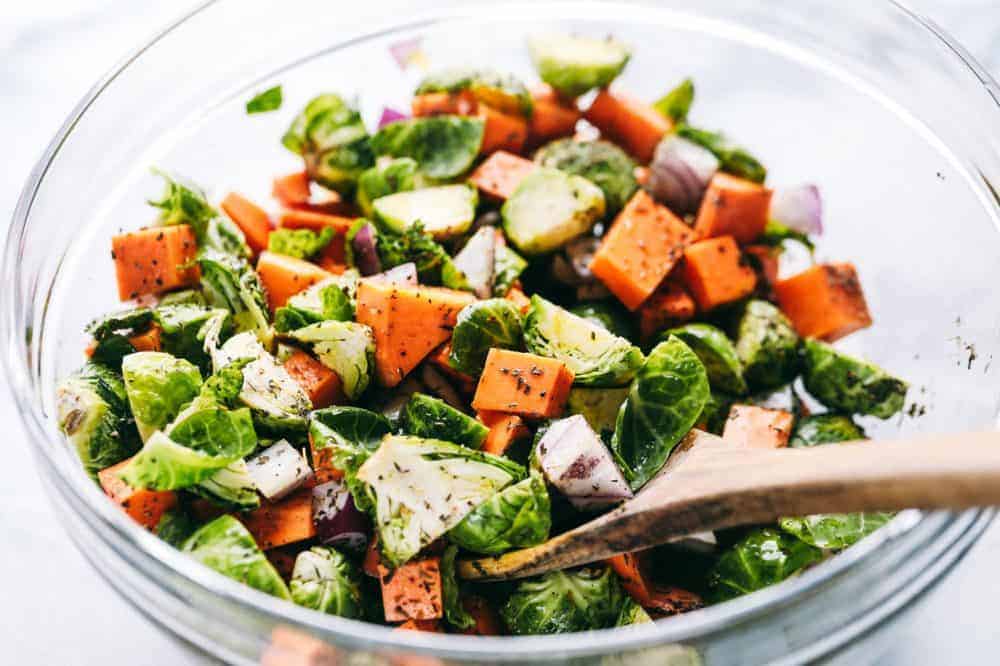 Chopped up carrots, brussels sprouts and red onions being stirred together with seasoning from McCormick using a wooden spoon and a glass bowl.