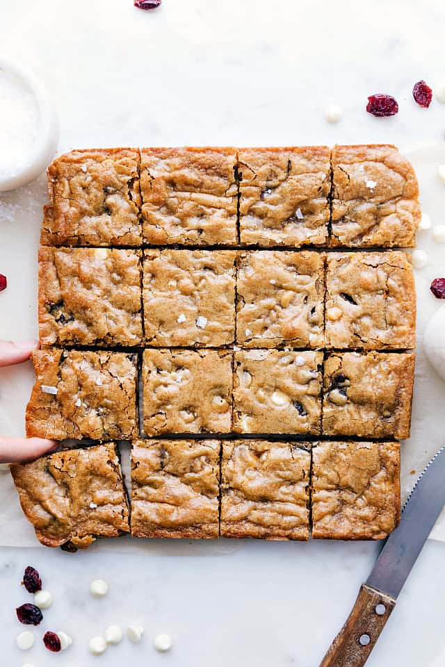 White chocolate cranberry Blondies cut into 1 inch squares makingIs 16 piecesWith a knife on the side and white chocolate chips and cranberries garnished around the blondies. 