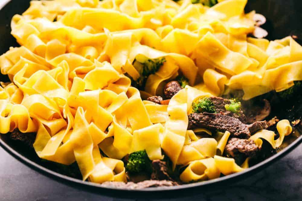 Thick egg noodles are being placed in the skillet with garlic beef and broccoli. 