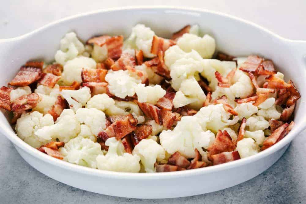 Chopped up cauliflower with Smithfield bacon pieces cut up in a white casserole dish being ready to bake.