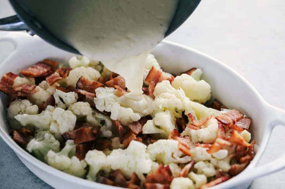 Chopped up a cauliflower and Smithfield bacon pieces in a white casserole dish having the au gratin being poured over top is it from the sauce pan.