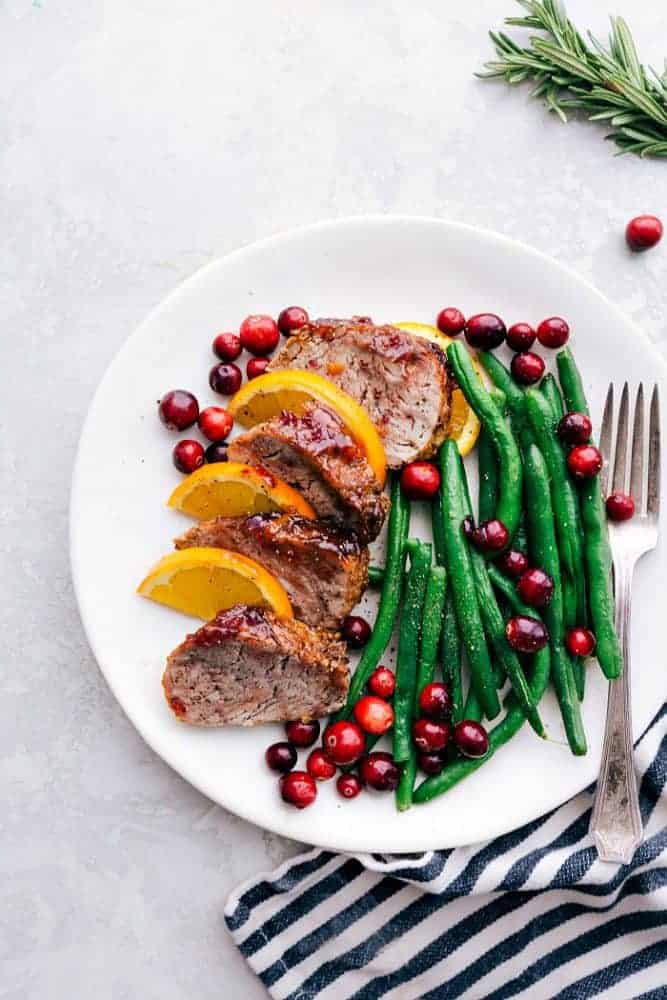 Roasted cranberry orange pork tenderloin with green beans on a white plate with a metal fork and a black and white cloth on the side.