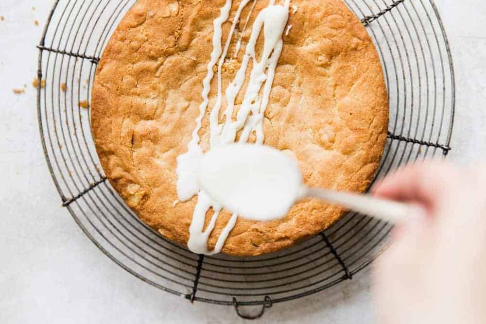 Spoon drizzling almond topping over brown butter almond cake.