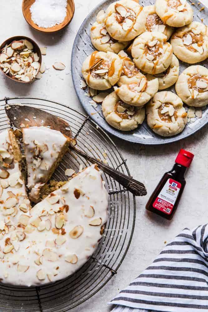 Brown butter almond cake on the cooling rack with a slice being removed. There is also a plate of brown butter almond cookies on the side. There is a small bottle of McCormick Almond Extract also. 