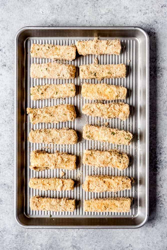 An image of breaded mozzarella sticks on a baking sheet for freezing.