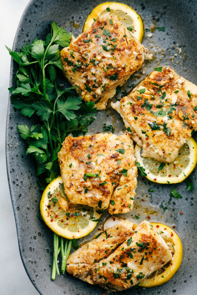 Baked cajun garlic butter cod served on a black speckled plate with green garnish and lemon slices underneath the cod filets. 