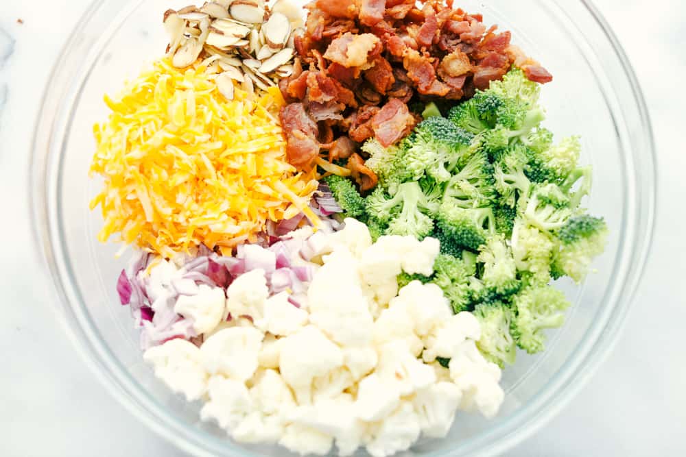 Shredded cheese, sliced almonds, bacon chopped up, broccoli and cauliflower florets and red onion chopped up separated in a glass bowl. 