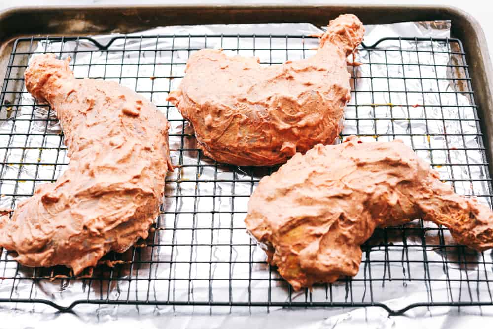 The marinated chicken sitting on a cookie rack with aluminum foil underneath on top of a baking sheet. 