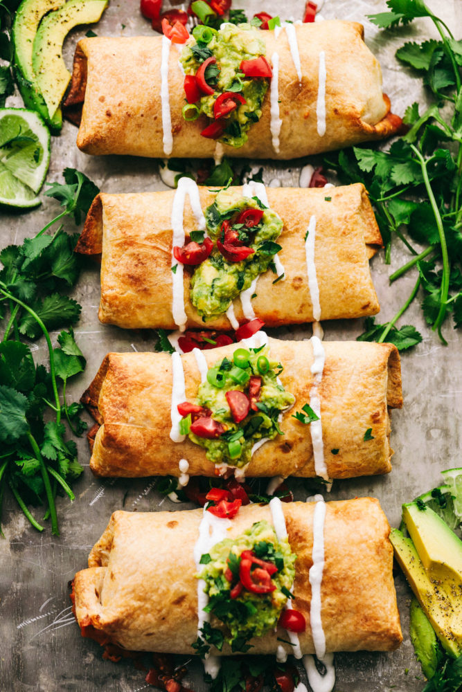 Four chicken enchilada lined up with guacamole, tomatoes and seasoning over top. 