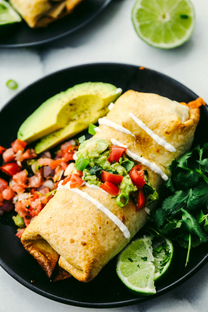 Chicken chimichanga on a black plate with an avocado sliced and limes slices on the side. 