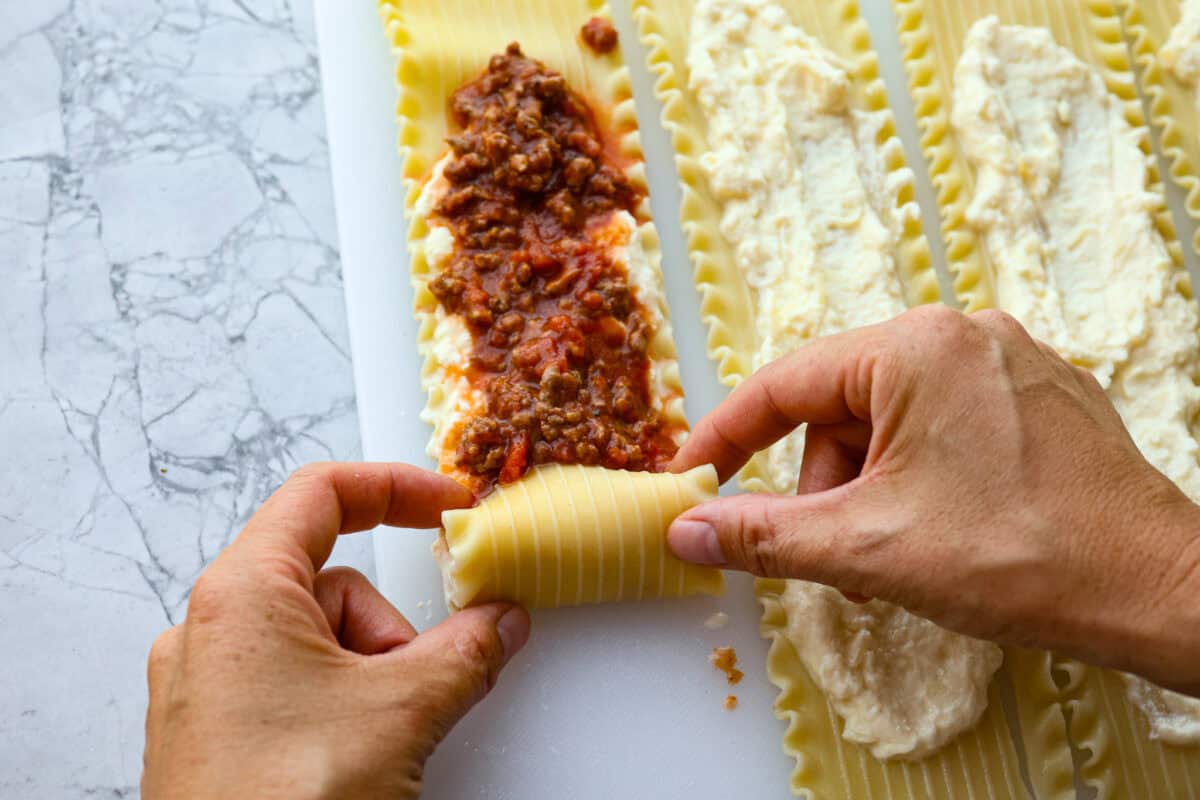 Overhead shot of someone rolling up the lasagna noodle with cheese and sauce spread on top. 