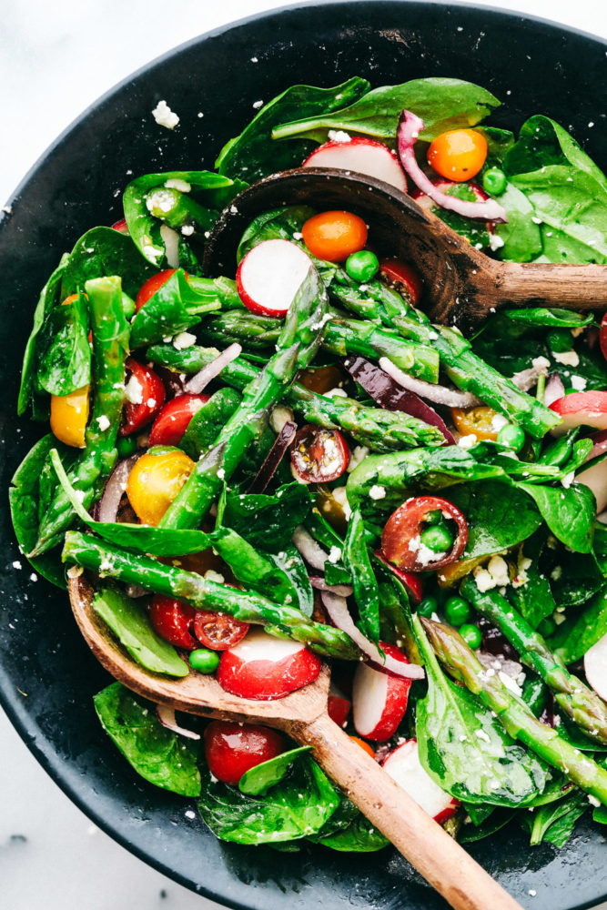 Asparagus salad being tossed together in a black bowl with two wooden spoons.