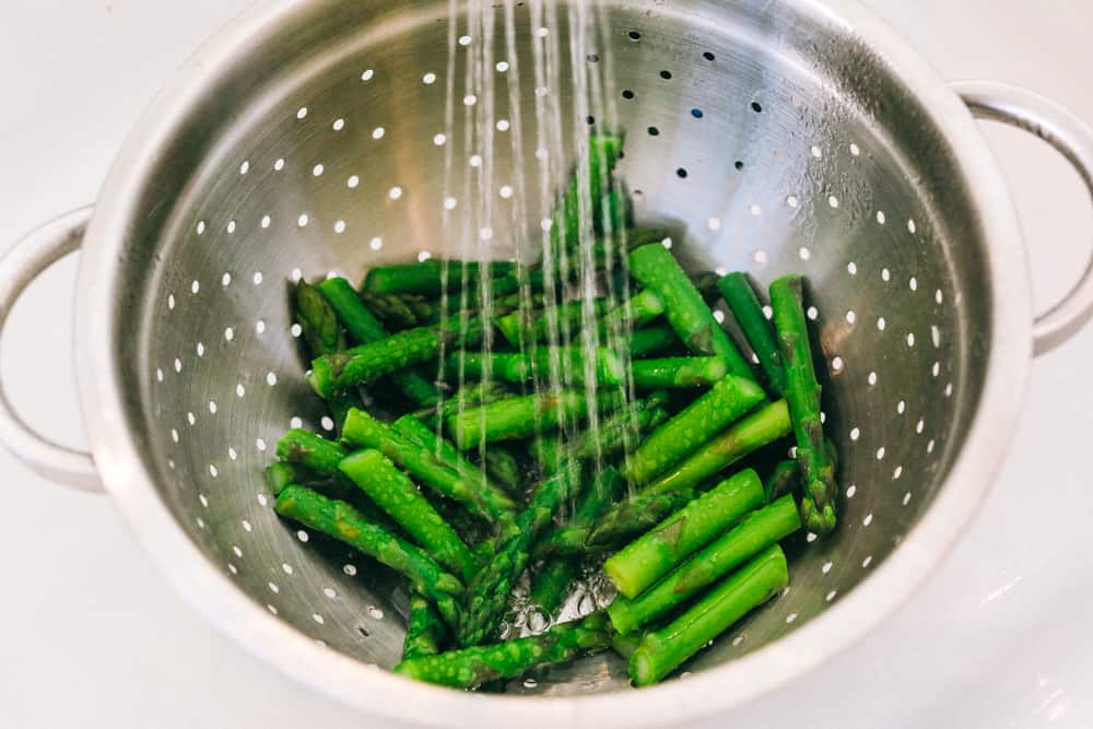 Asparagus being washed with cold water in a strainer. 