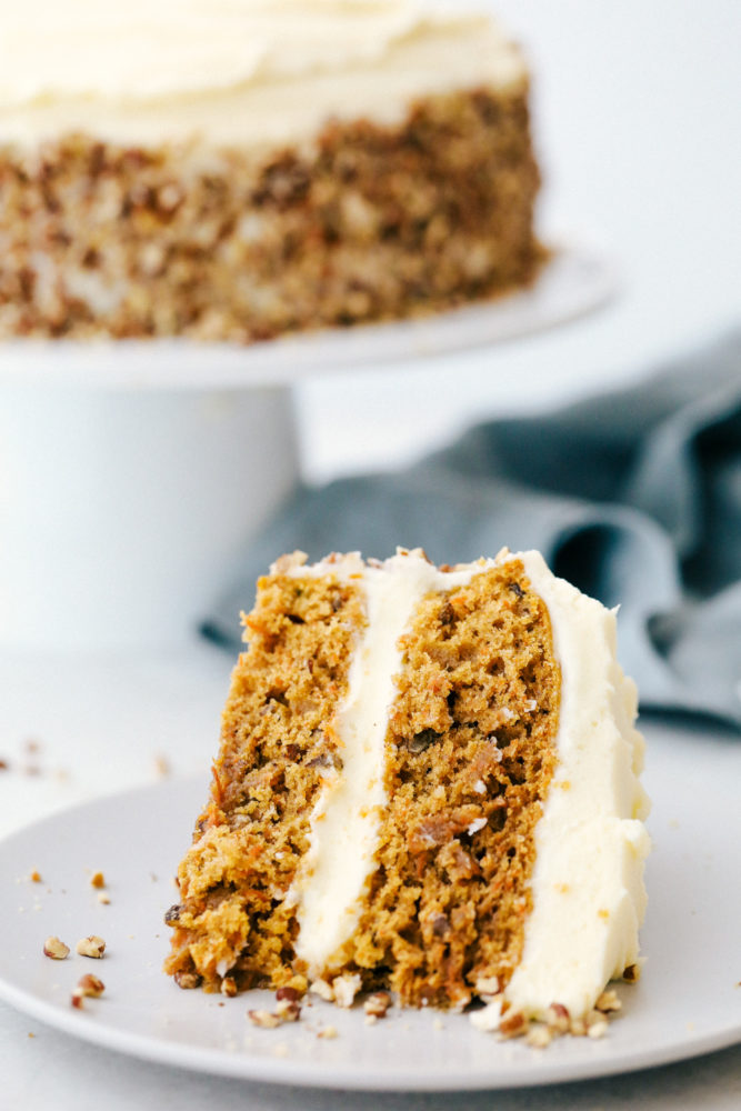 A slice of carrot cake on a plate with the carrot cake in the background on a cake platter. 