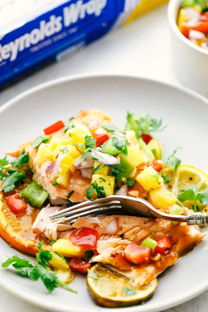 Grilled citrus salmon on a plate with pineapple salsa over top and being cut into with a silver fork. 