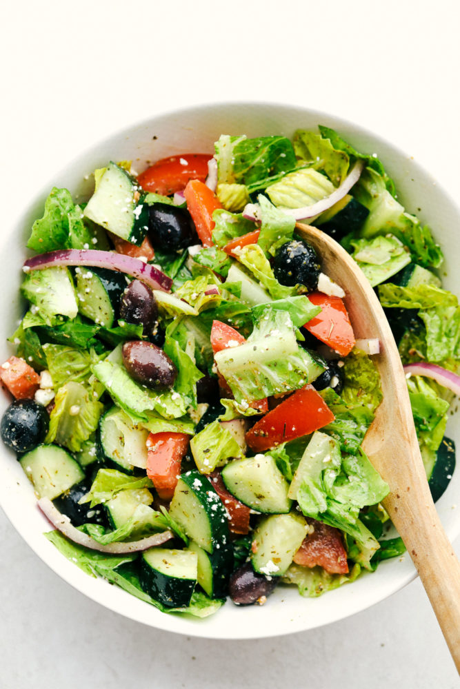 Greek salad in a white bowl with a wooden spoon.