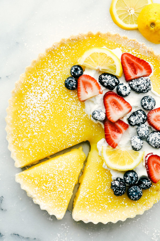 Lemon tart photo with fresh berries on top with lemon slices and powder sugar sprinkled on top and a slice cut out of the tart. 