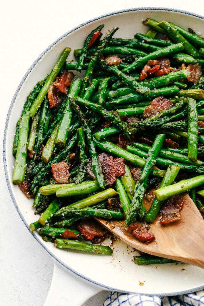 Garlic asparagus with bacon being stirred in a skillet with a wooden spoon. 