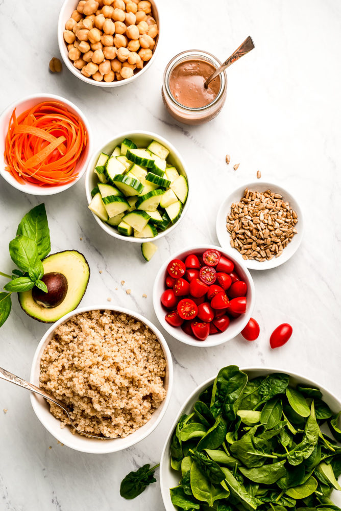 Various size bowls of vegetables, quinoa, seeds, chickpeas, and dressing.