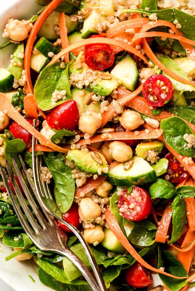 A salad of spinach, cucumbers, grape tomatoes, carrot ribbons, avocado, chickpeas, and quinoa all mixed together.