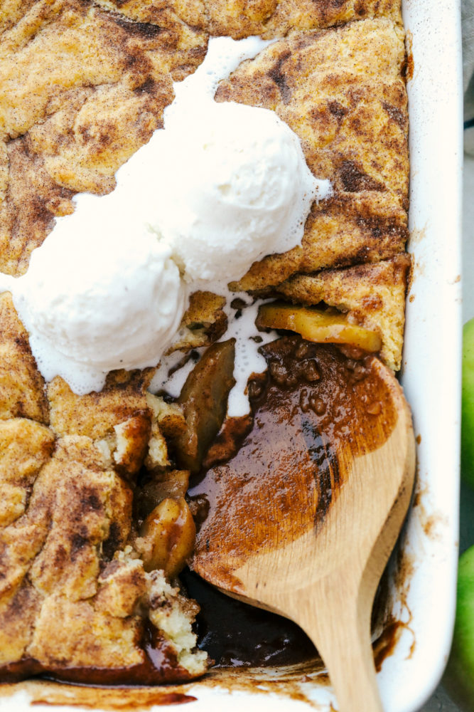 Scooping apple cobbler and vanilla ice cream out of a white baking dish.