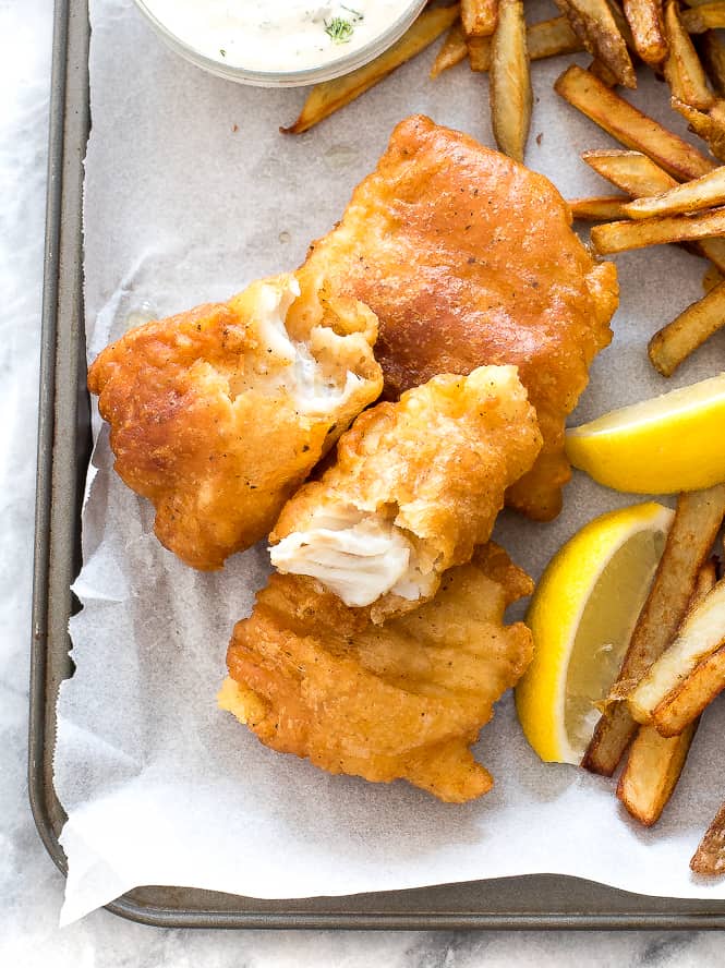 Fish and chips on a sheet pan with parchment paper overtop with French fries and lemon slices on the side. 