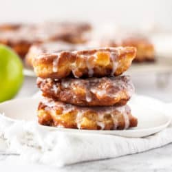 A stack of 3 apple fritters on a white plate with more in the background