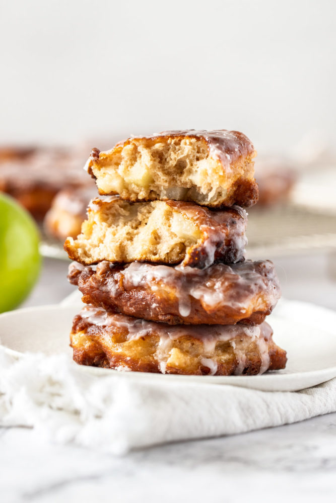 A stack of apple fritters on a white plate, the top one is broken in half showing the inside.