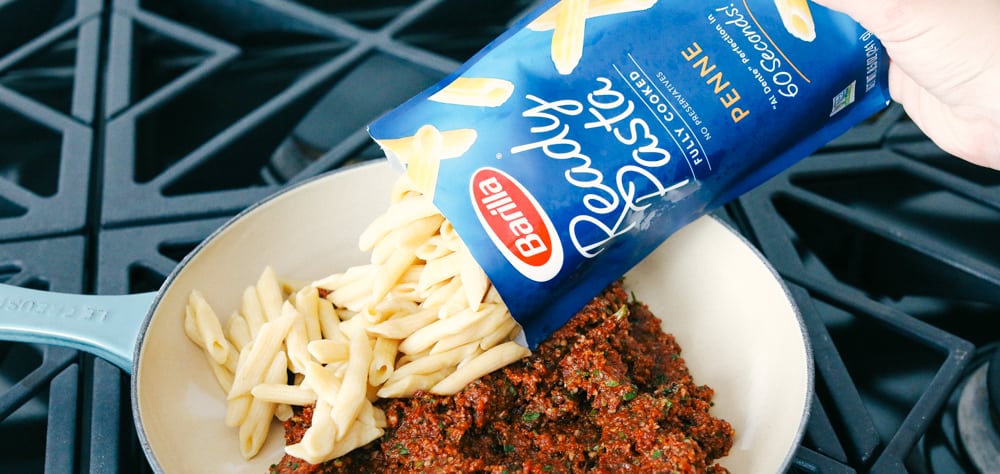 Cooked ready pasta being poured in the skillet with the sun dried tomato pesto sauce. 