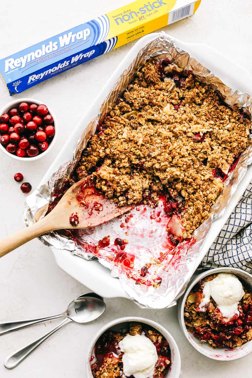 Cranberry apple crisp in a white casserole dish with cranberries and Reynolds wrap on the side. 