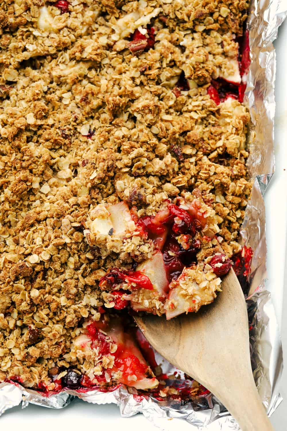 Cranberry apple crisp in a baking pan with a wooden spoon taking out a scoop. 