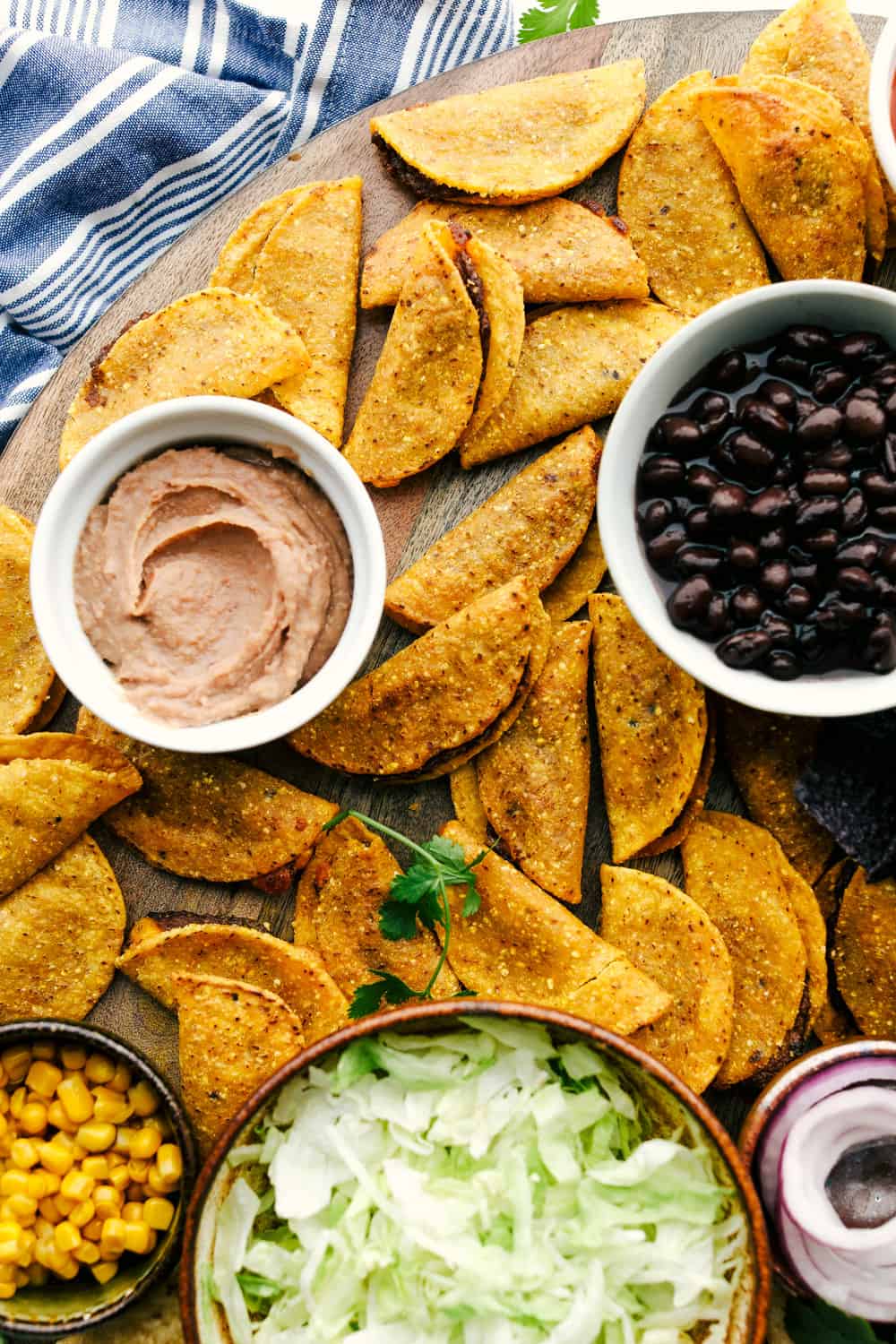 Taco board with tacos, bean dip, black beans, corn and lettuce in bowls on a board.