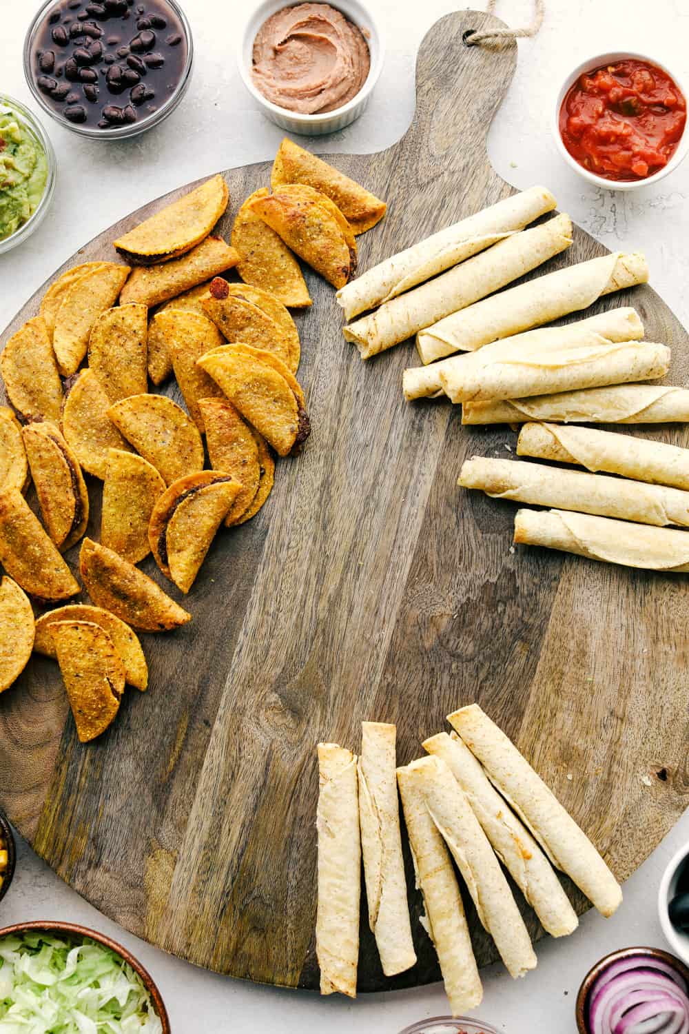 A board with tacos and taquitos on it with bowls of dips and salsa around the edge. 