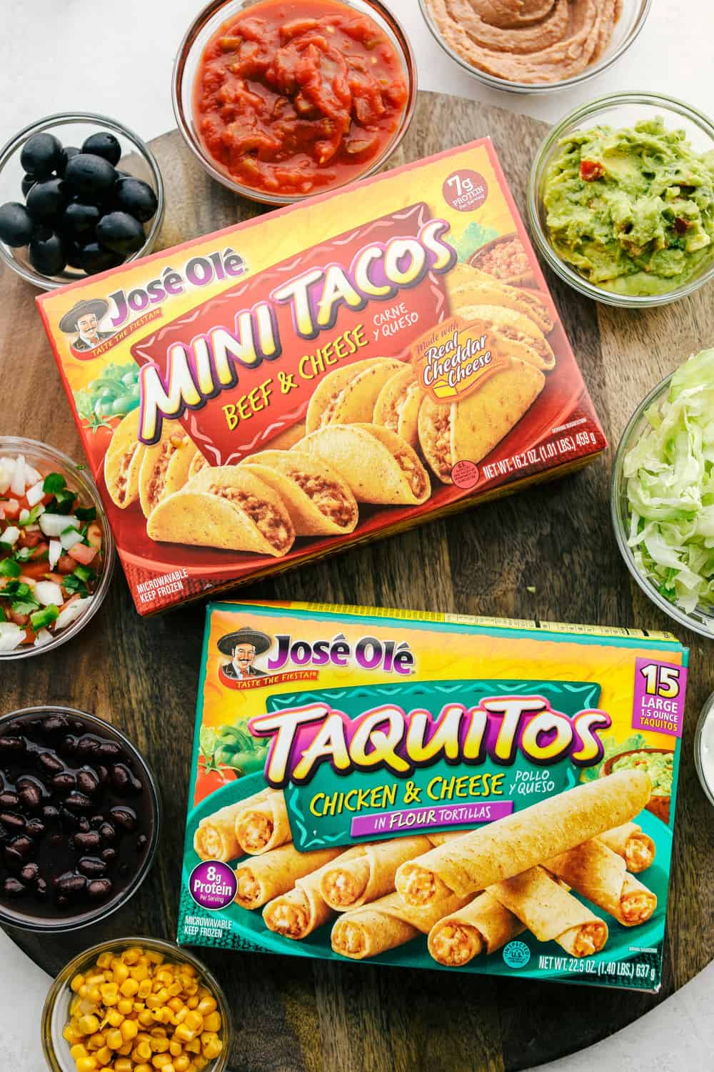 Mini tacos and taquitos in a box with bowls of dips and vegetables. 