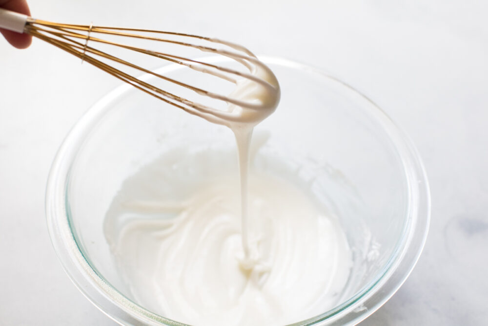 Whisk dripping with frosting over a glass bowl. 