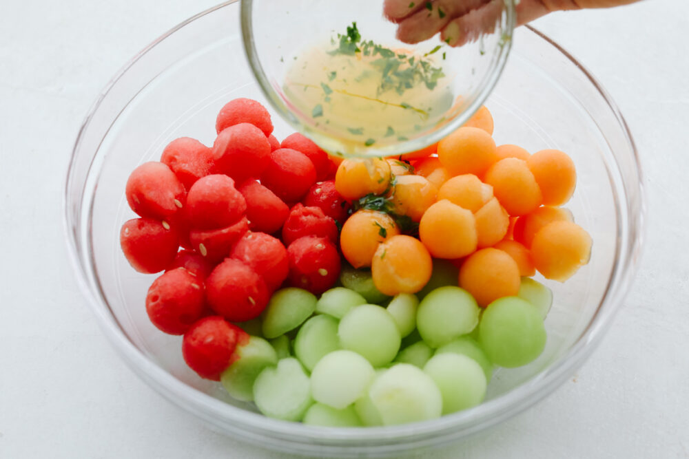 A bowl filled with watermelon, honeydew and cantaloupe scooped in small sizes with dressing being poured on them. 