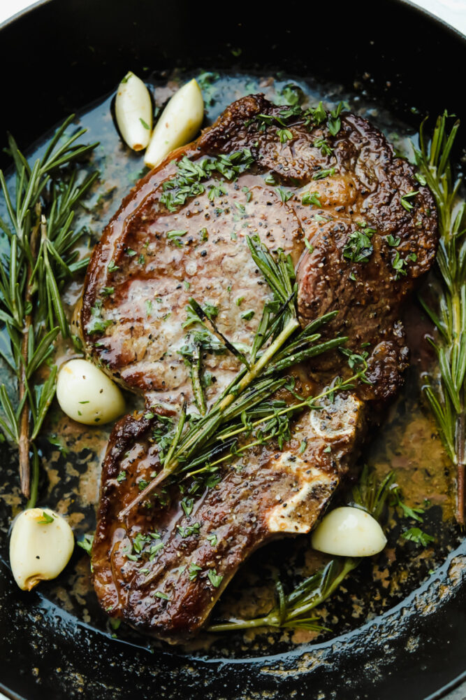 Cooked ribeye steak garnished with rosemary, thyme, and garlic.