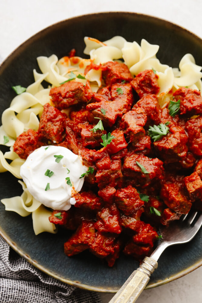 A bowl of pörkölt on top of wide egg noodles with a dollop of sour cream on top. 