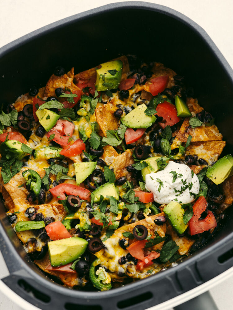 An air fryer loaded with nachos topped with cheese, vegetables, sour cream and cilantro.