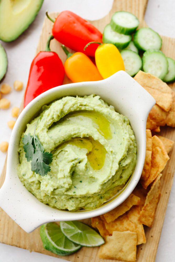 Avocado hummus in a bowl on a wooden cutting board with some pita chips, cucumbers and peppers. 