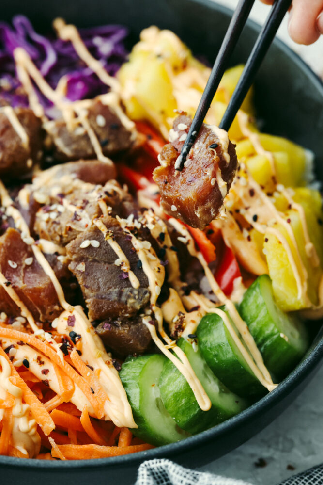 A hand holding chopsticks with a piece of tuna coming out of a Hawaiian poke bowl 