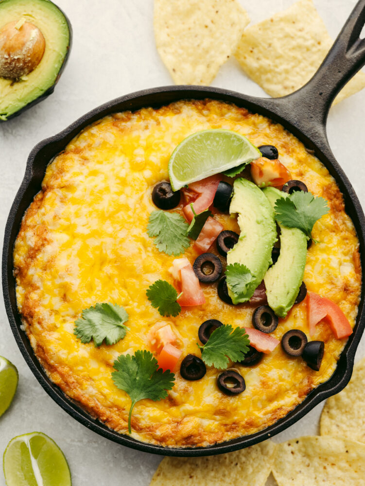 A pan of Texas trash dip garnished with olives, cilantro, a lime wedges and some tomatoes and avocado. 