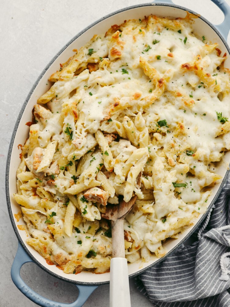 A baking pan filled with chicken and noodles and sauce with a wooden spoon. 