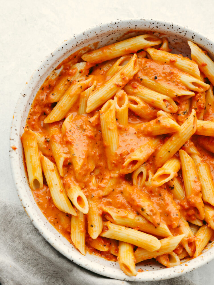 Penne coated with red pepper pasta sauce in a white speckled stoneware bowl.