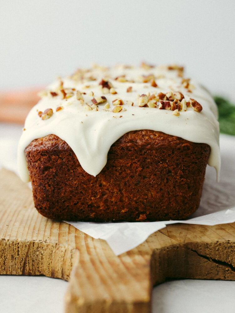 A side view of a loaf of bread sitting on a wooden cutting baord with a glaze and nuts on top. 