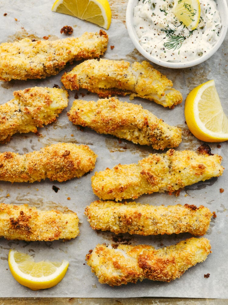 Baked cod fish sticks on a baking sheet with a slice of lemon and some tarter sauce in a small bowl. 