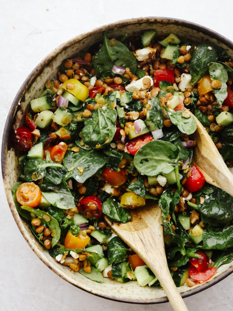 Tossing lentil salad with 2 wooden spoons in a brown bowl.