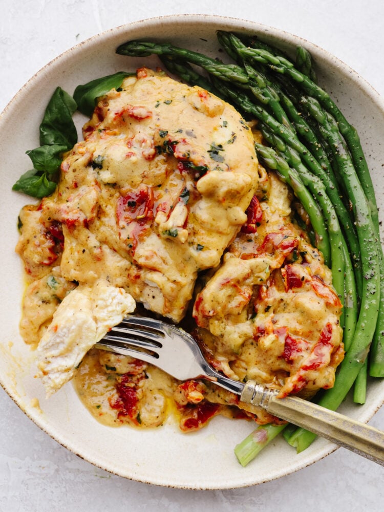 A plate with chicken and asparagus. A silver fork is cutting into the chicken. 